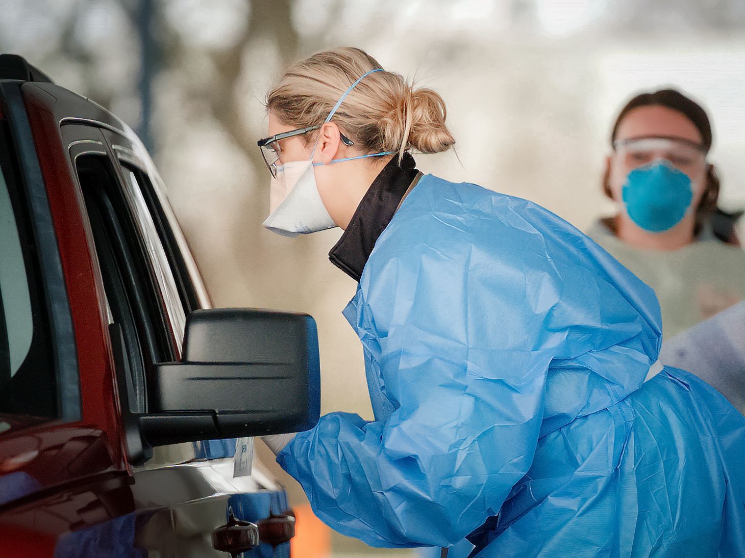 nurse in personal protective equipment speaking to someone through a car window at a drive through coronavirus testing site