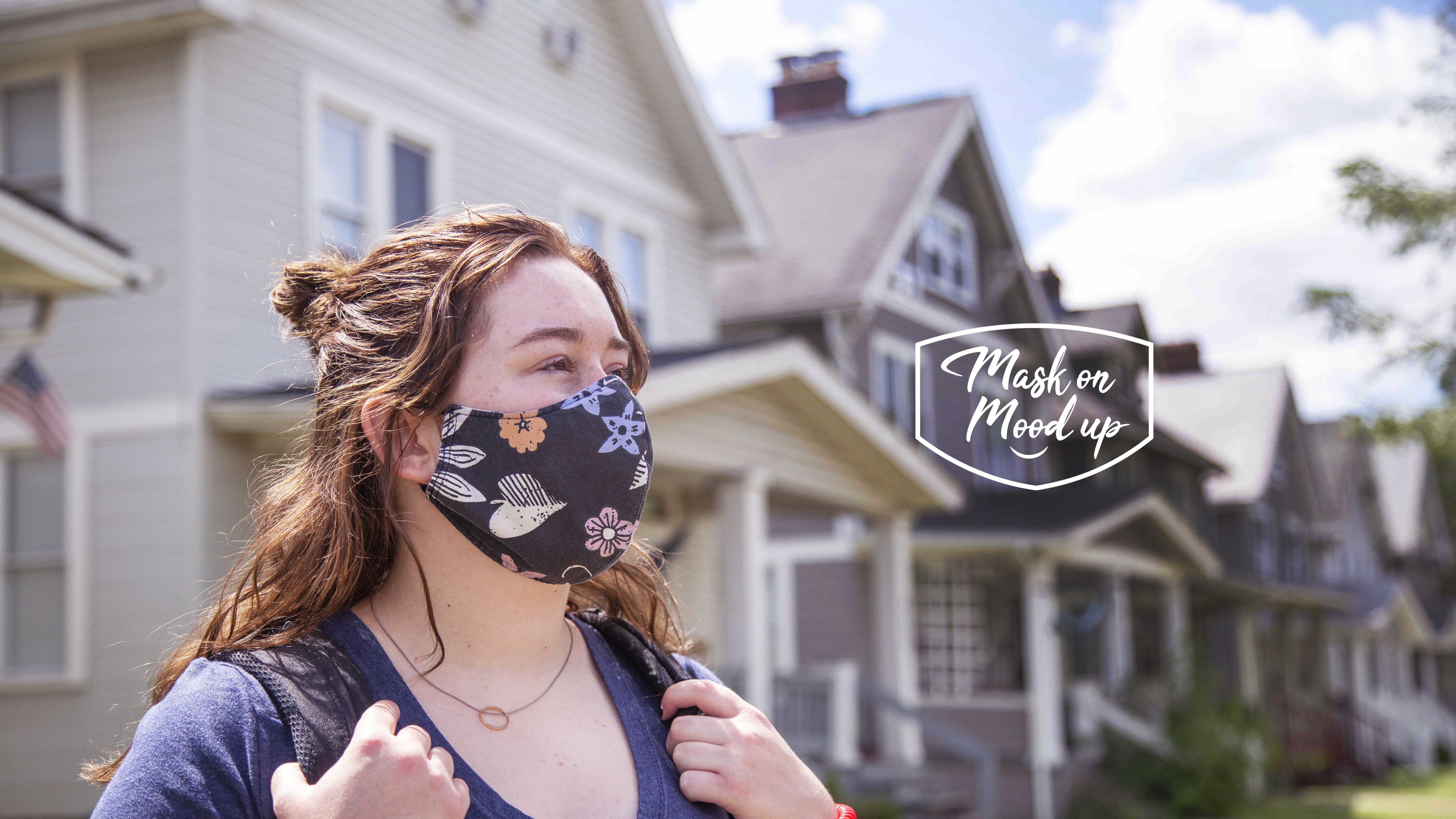 female student wearing backpack and mask in front of houses