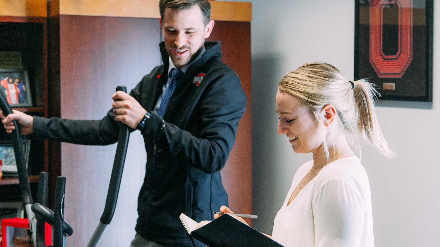 instructor exercising on elliptical and student taking notes