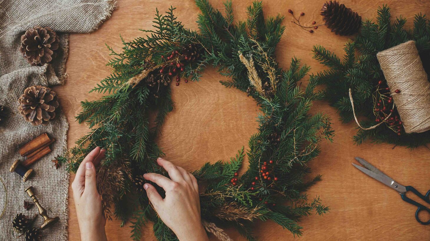 Hands working on a holiday wreath at a table with craft supplies