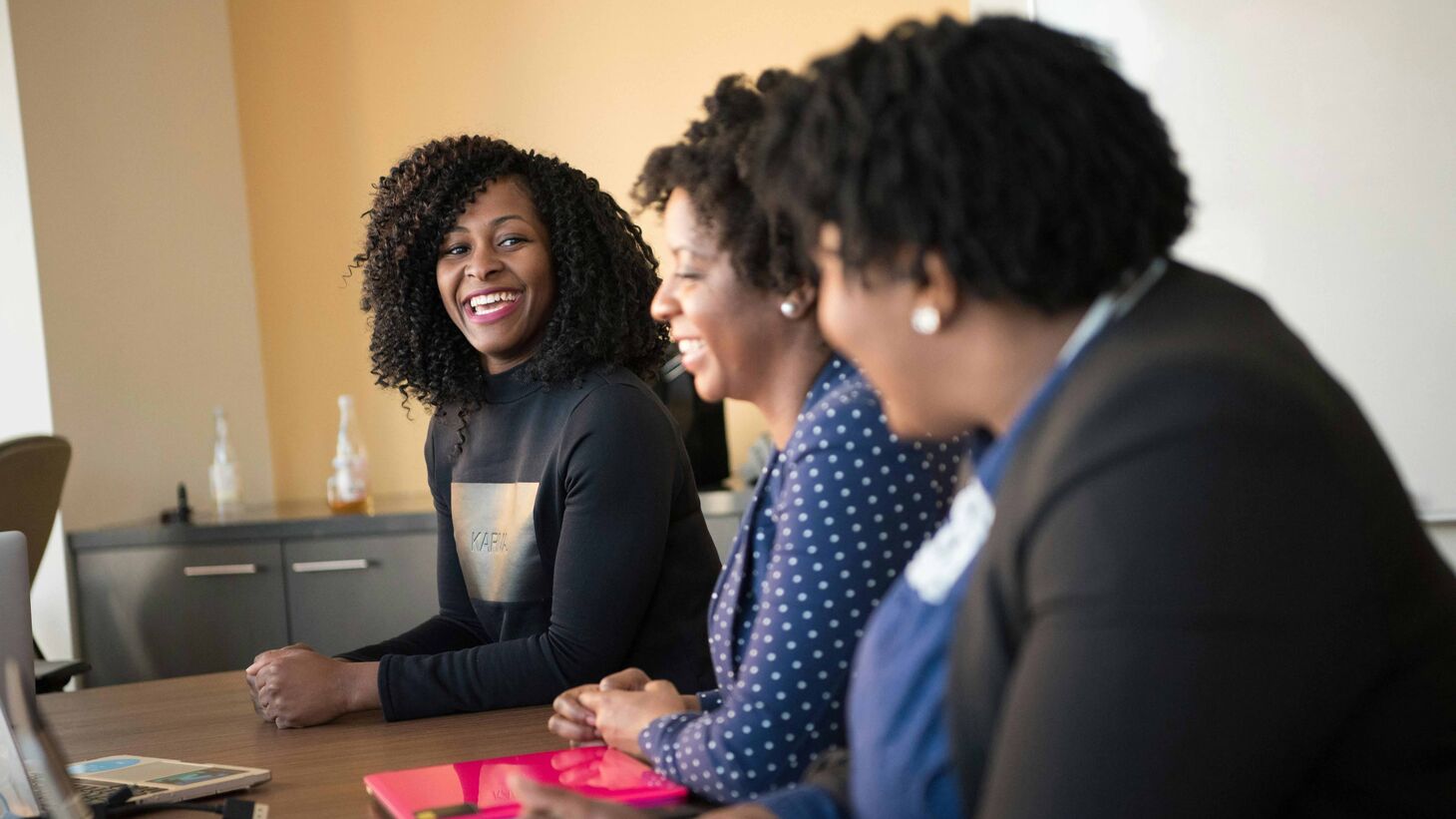smiling woman leaning on desk while talking to two other women