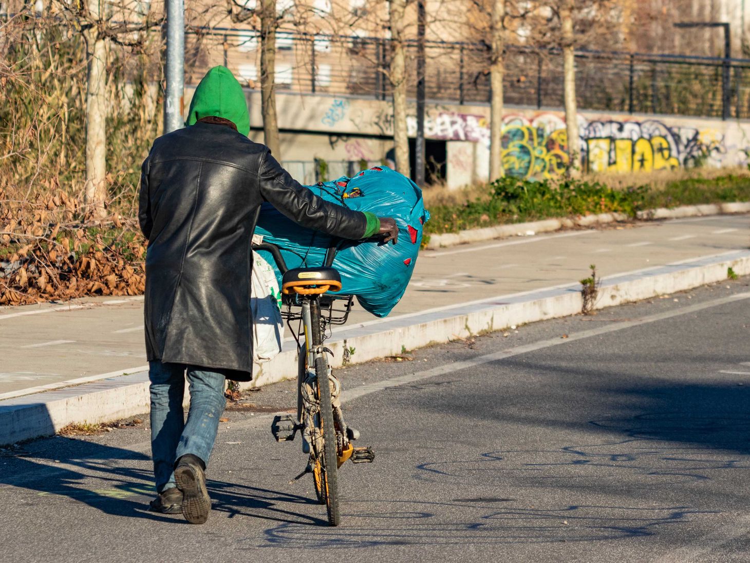 back of homeless man pushing a bicycle with a bag of belongings balanced on top