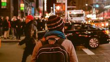 young woman wearing backpack and winter hat waiting to cross busy street in a city