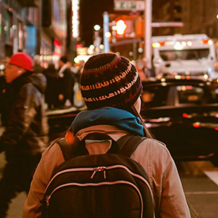 young woman wearing backpack and winter hat waiting to cross busy street in a city